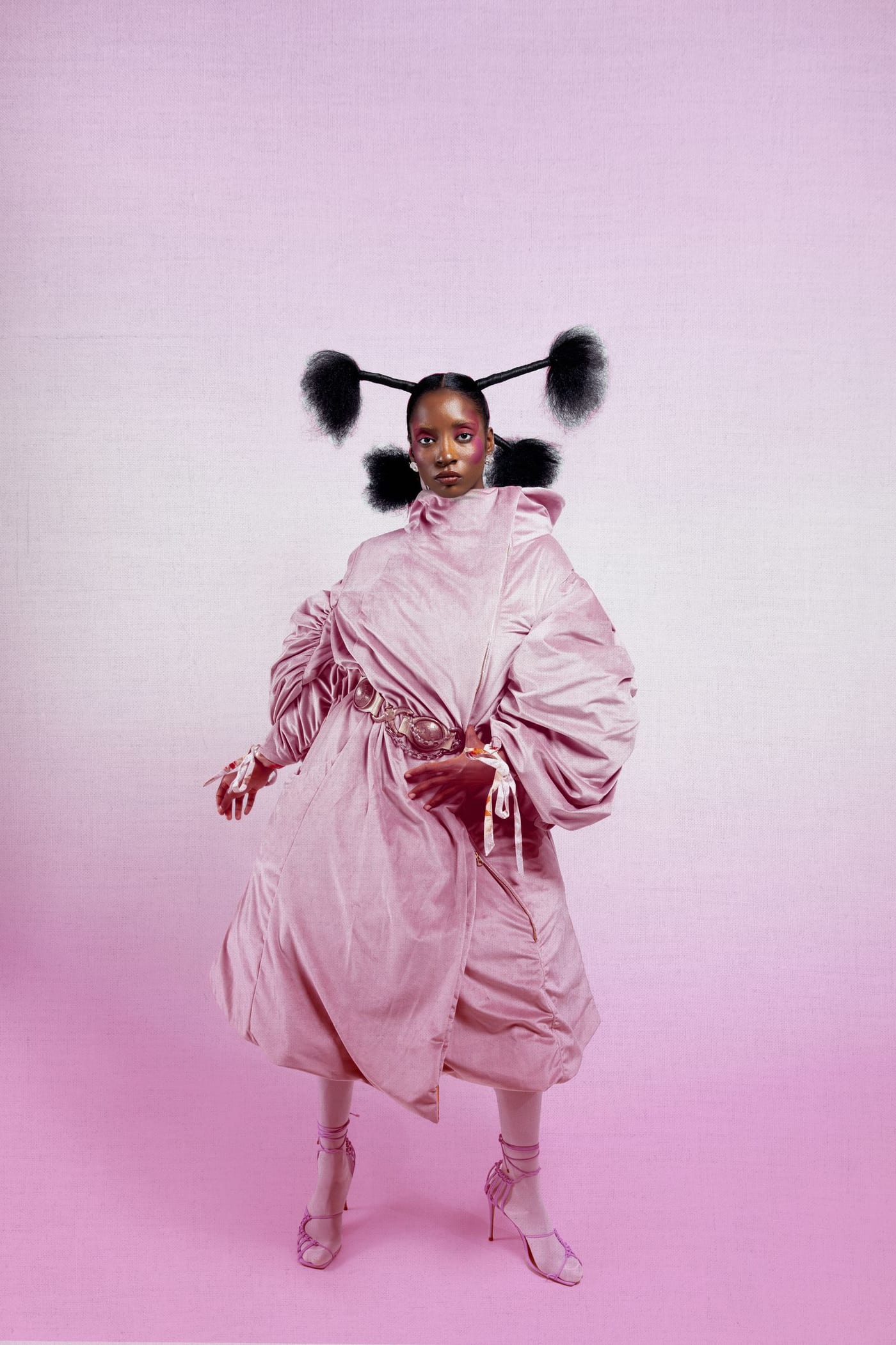 Woman in a sculptural blush velvet coat with gathered sleeves and pink strappy heels against a pink backdrop