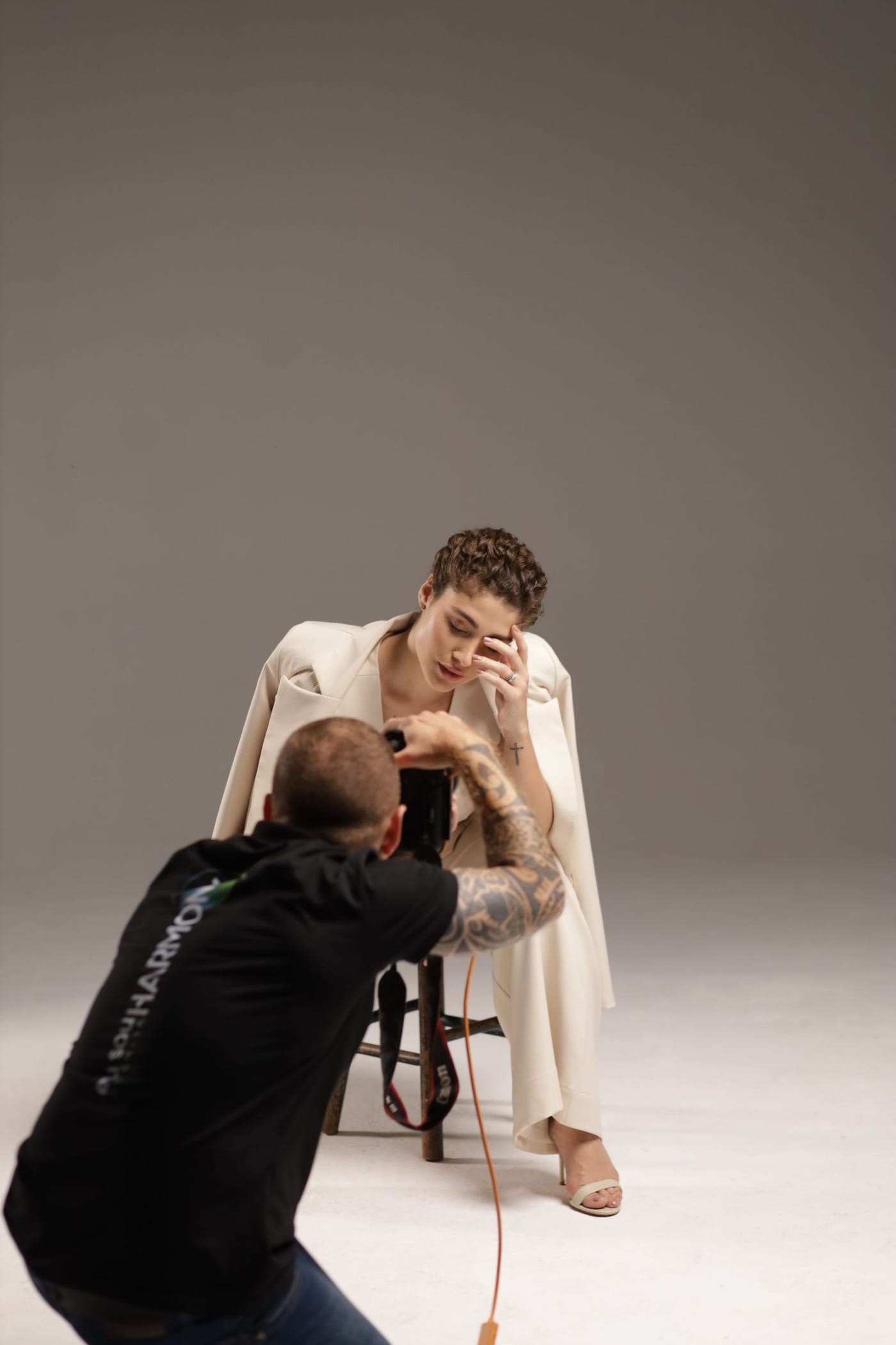 Woman in a cream suit with the jacket over her shoulders and nude sandals, sitting on a stool while a photographer shoots her