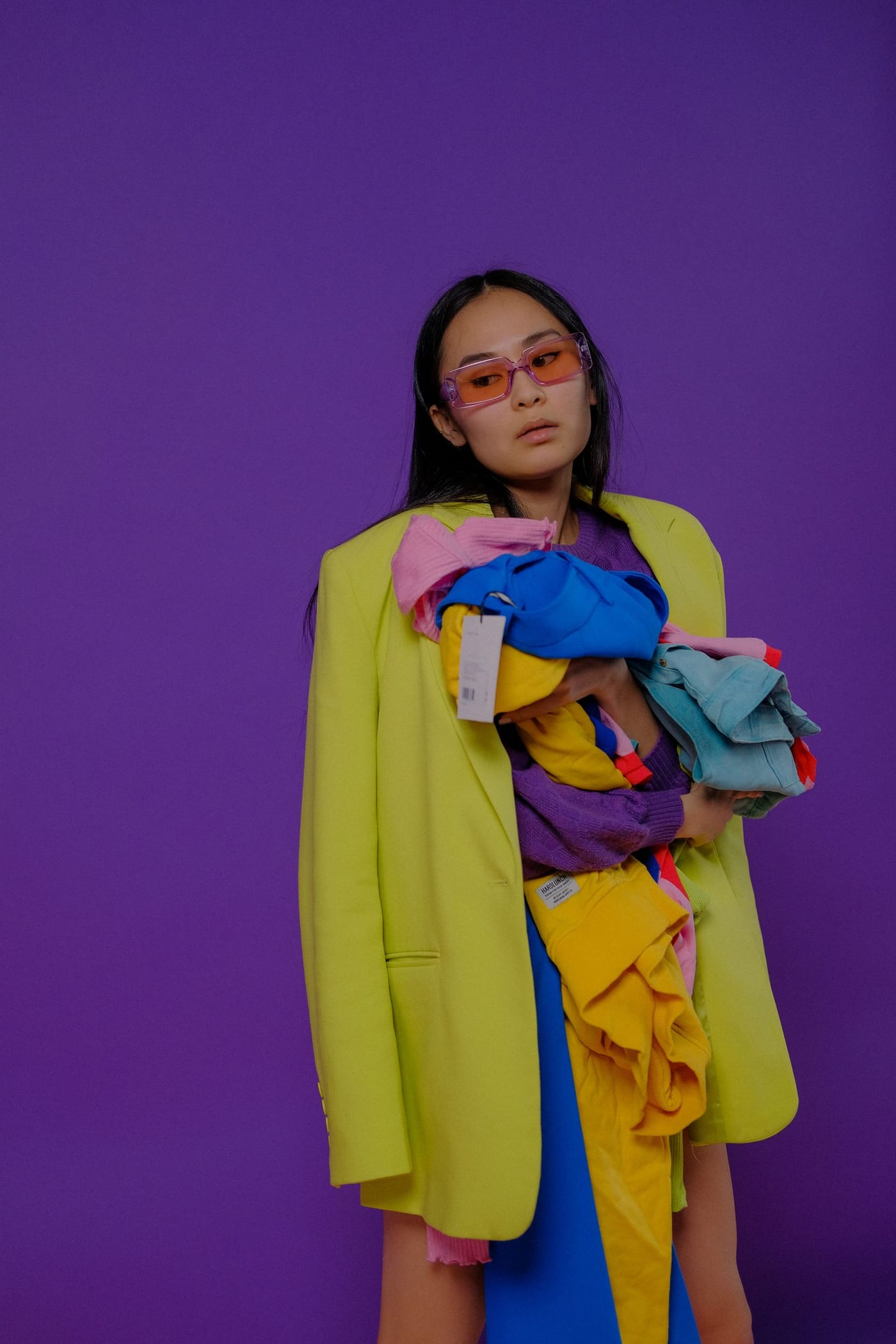 Woman in an oversized lime blazer and pink tinted glasses holding an armful of bright clothes against a purple wall