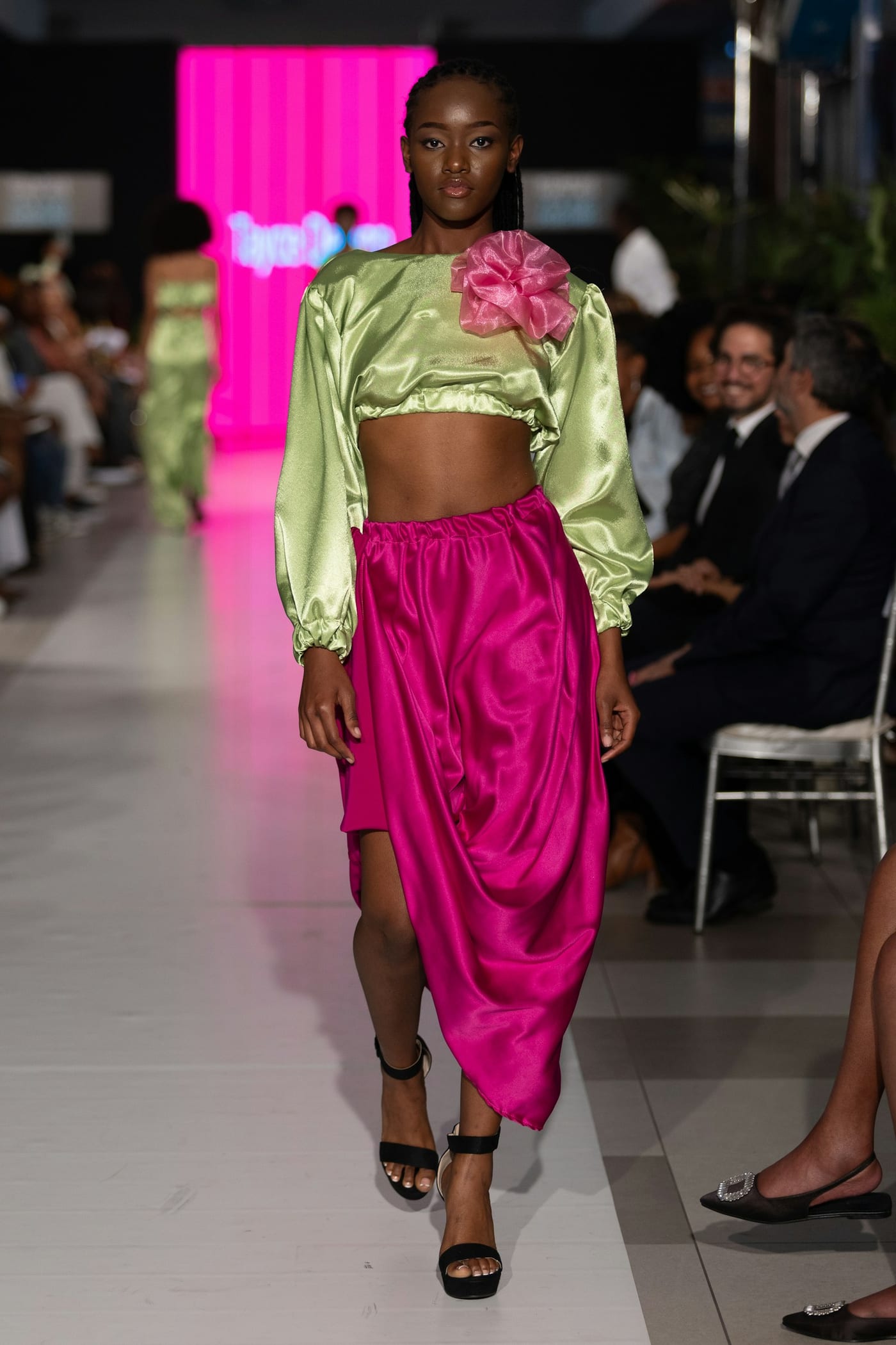 Woman on a runway in a lime satin cropped blouse with a pink organza flower and a draped fuchsia satin skirt